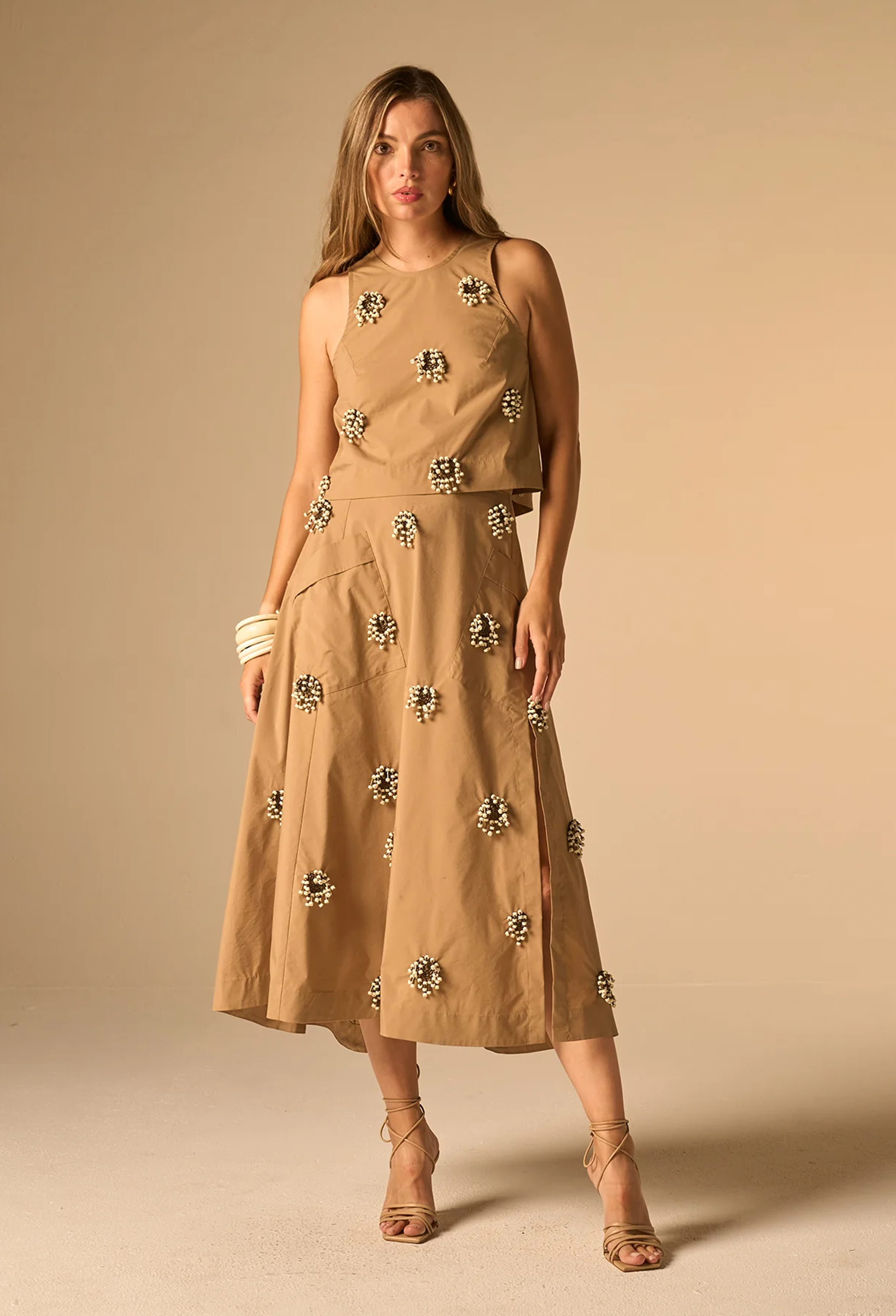 A woman models a sleeveless tan midi dress with a matching belt and scattered metallic floral embellishments, set against a solid beige studio background.