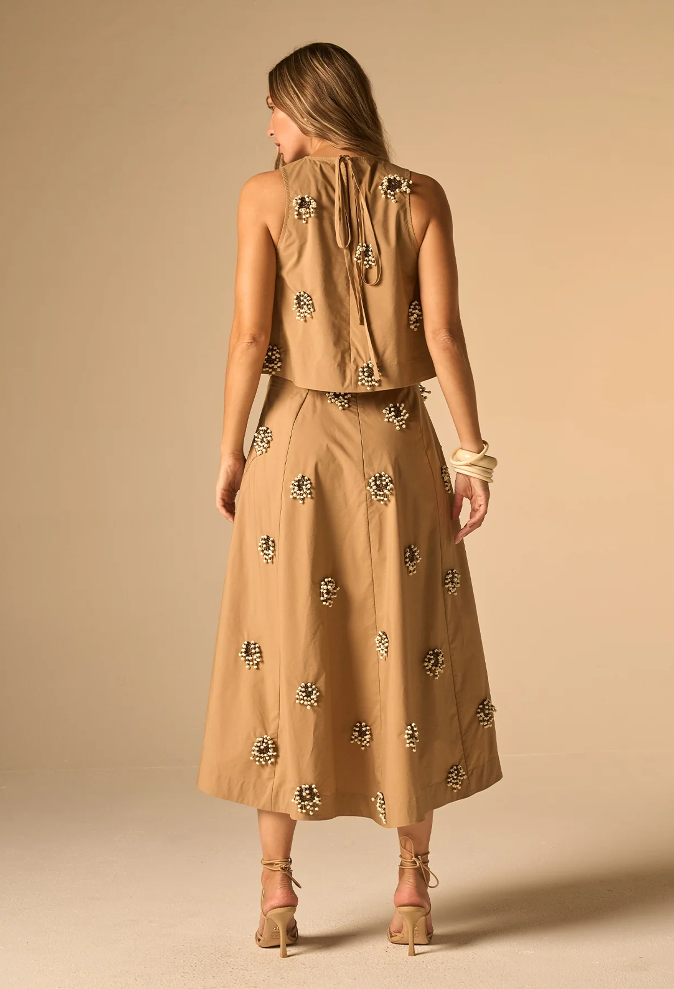 A woman shown from the back wearing a sleeveless tan midi dress with a tie-neck detail and scattered floral applique, paired with beige high-heeled sandals against a solid beige studio background.