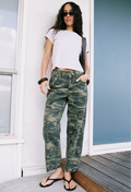 A woman stands outdoors wearing a white cropped T-shirt, camouflage cargo-style jeans, black flip-flops, and black sunglasses, with a dark shoulder bag visible.