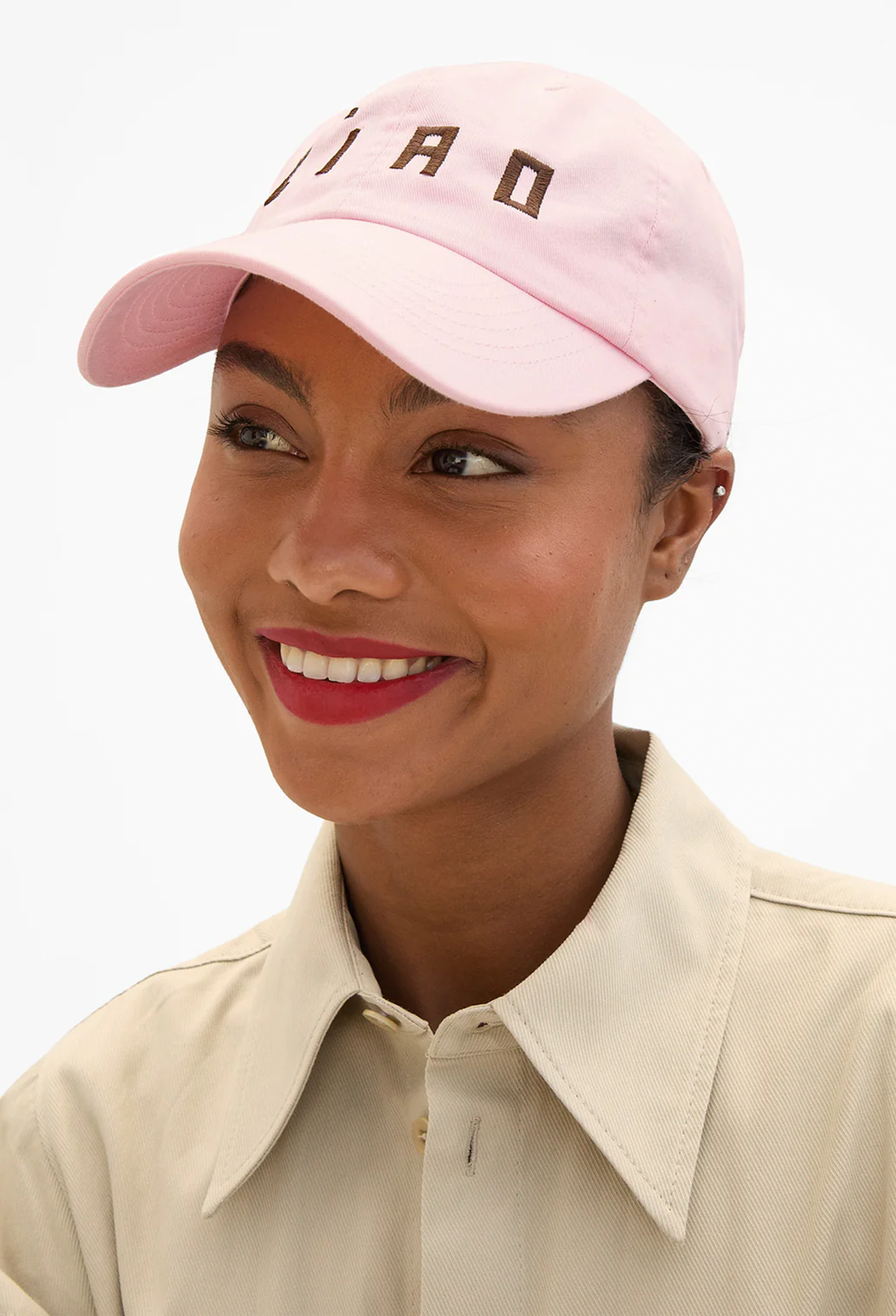 A smiling woman wearing a light pink baseball cap with dark brown embroidered letters and a cream-colored collared shirt, photographed against a clean white background.
