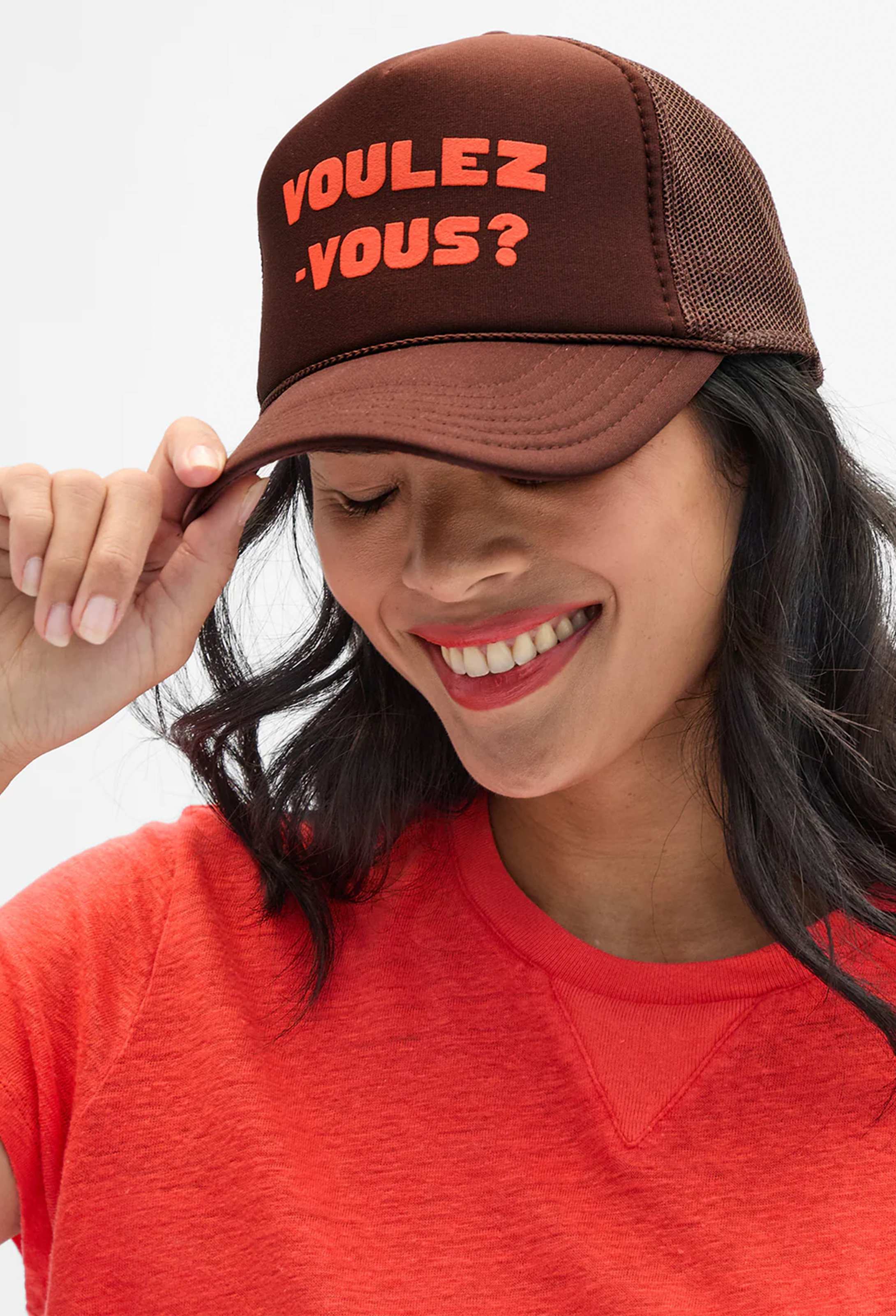 A woman wearing a brown trucker hat with orange text reading 'VOULEZ-VOUS?' and a bright orange-red t-shirt, photographed against a clean light gray background.