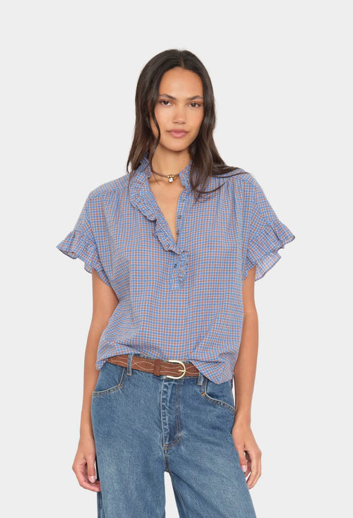 A woman modeling a short-sleeve button-front blouse with a ruffled collar and flutter sleeves in a blue gingham pattern, paired with blue jeans and a brown belt, against a light gray studio background.