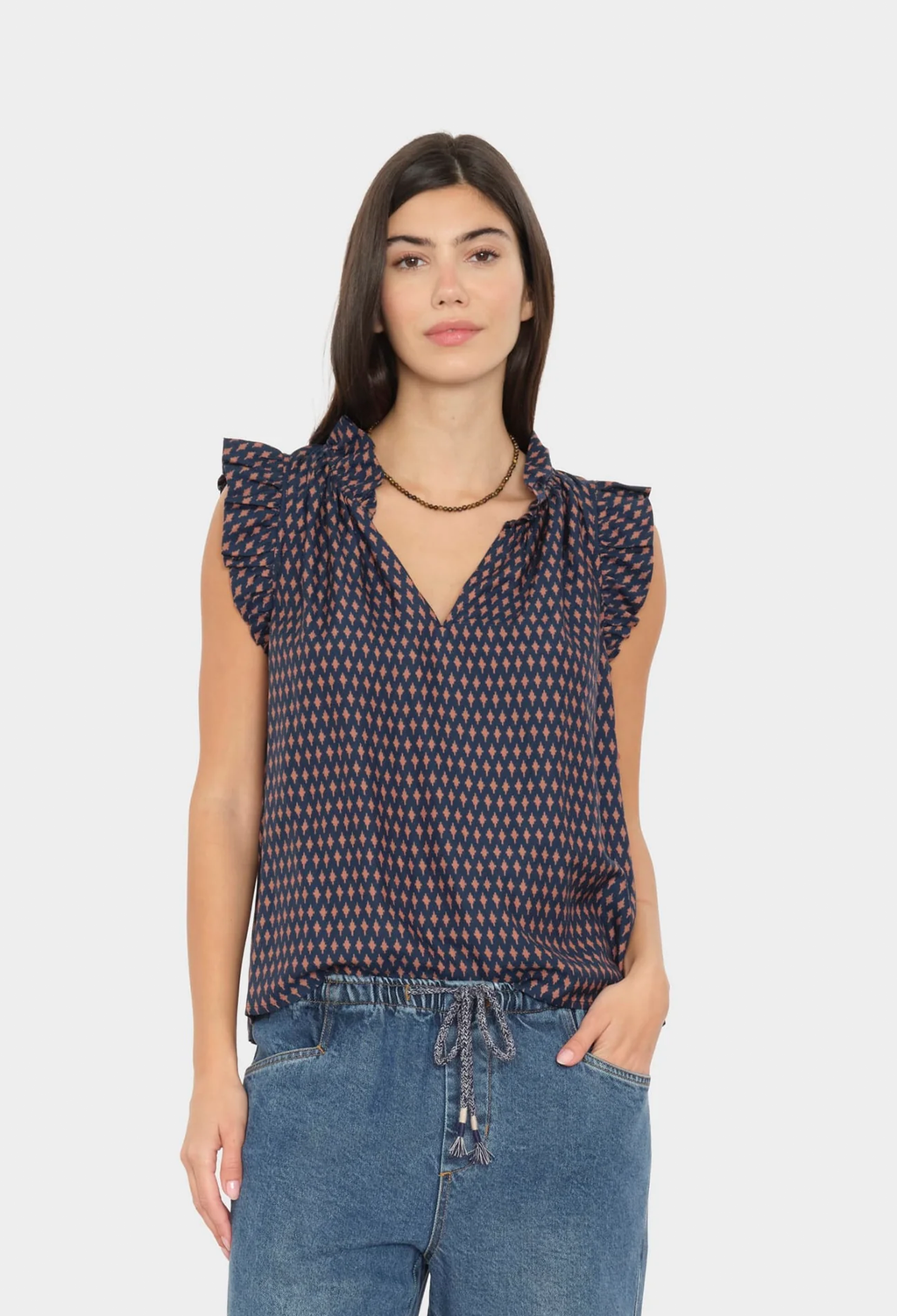 A woman modeling a sleeveless navy blue blouse with a small red and white geometric pattern, ruffled shoulders, and a tie-neck detail, paired with blue jeans against a light gray background.