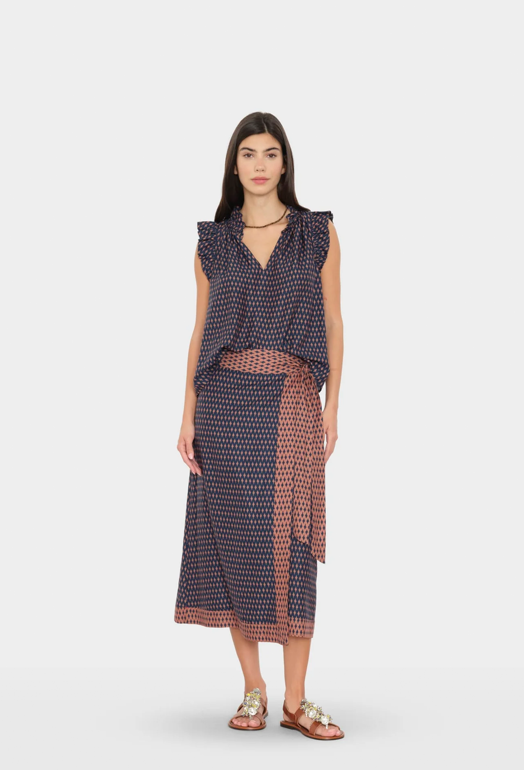 A woman modeling a sleeveless wrap-style midi dress with a navy and rust red geometric pattern, set against a plain light gray studio background.