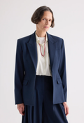 A model wearing a navy blue tailored blazer over a cream pleated shirt, paired with matching navy trousers and a red necklace, shot against a light gray studio background.