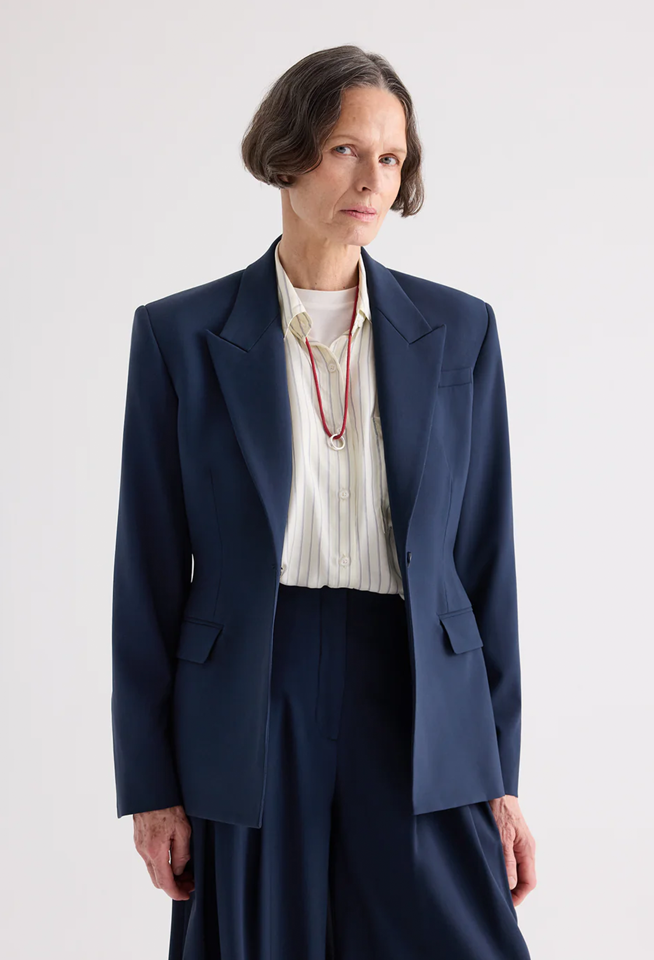 A model wearing a navy blue tailored blazer over a cream pleated shirt, paired with matching navy trousers and a red necklace, shot against a light gray studio background.