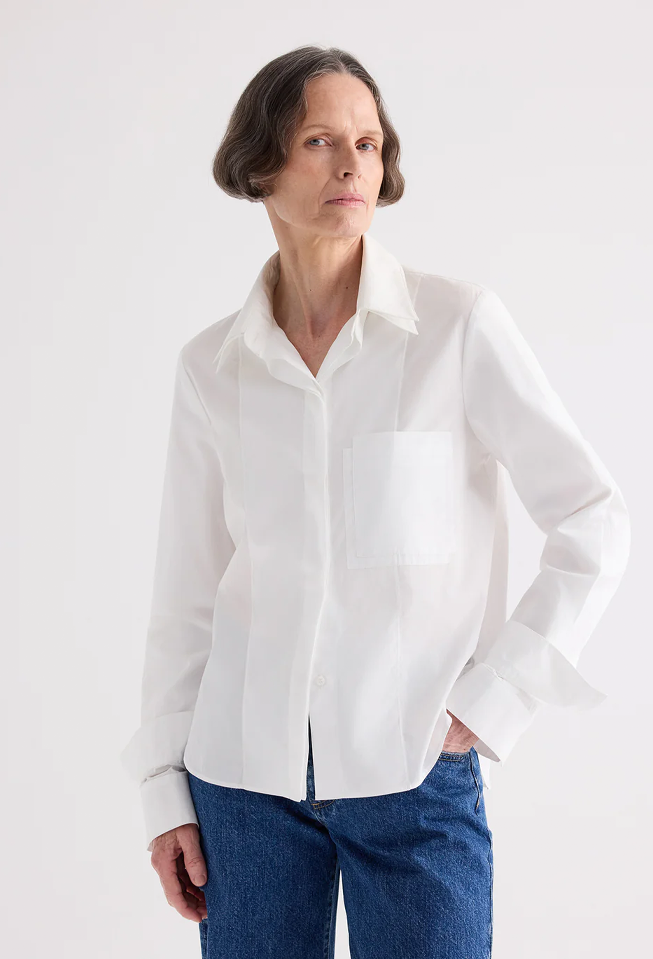 A woman wearing a white long-sleeve button-up shirt with a chest pocket and blue jeans, standing against a plain light gray studio background.
