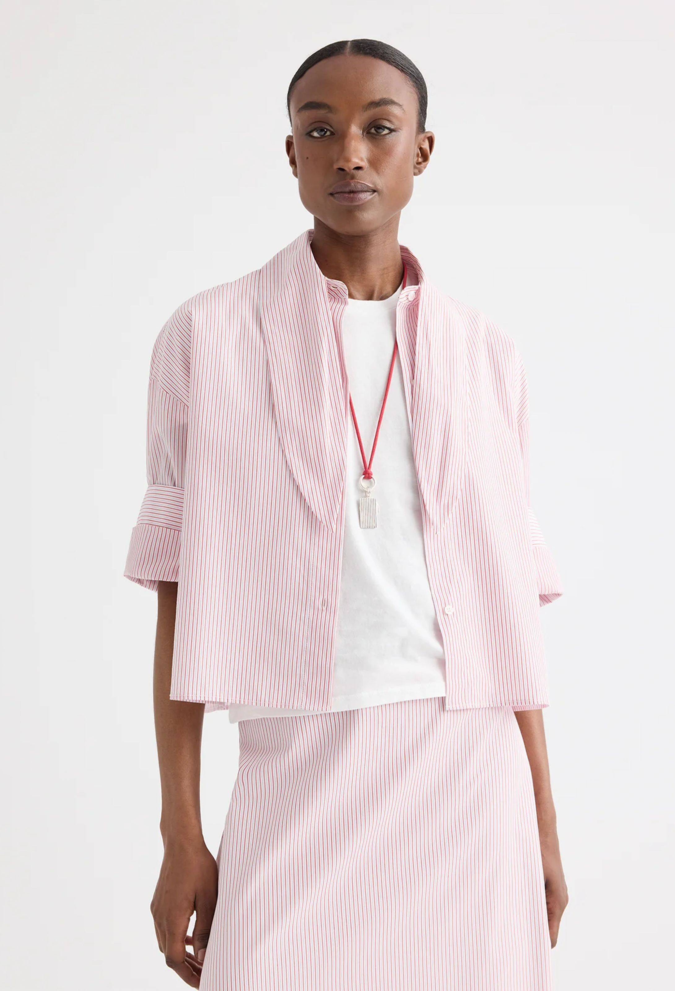 A model wearing a red and white vertically striped cropped shirt with a tie neck, paired with a white t-shirt and a matching skirt, set against a plain white studio background.