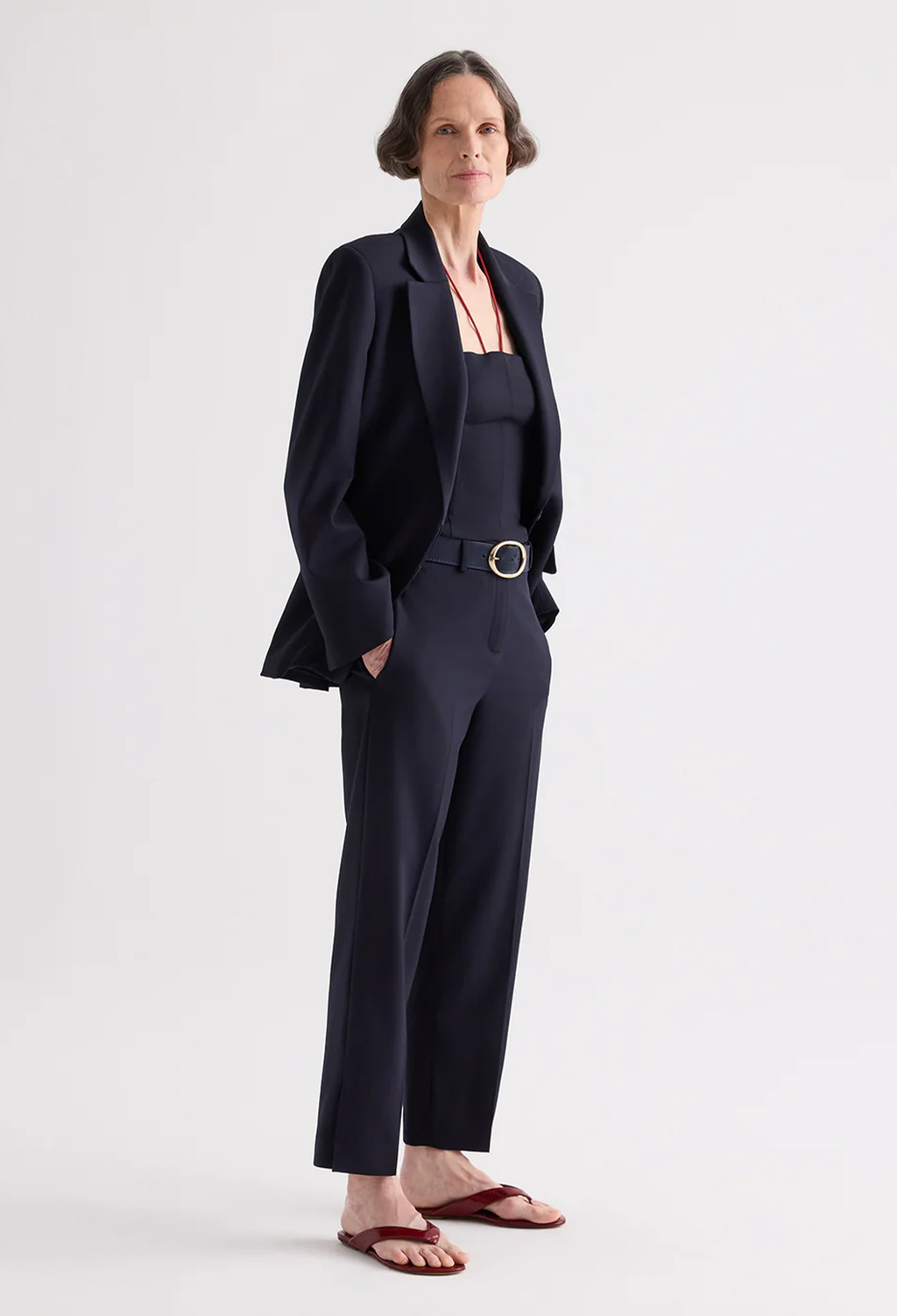A woman modeling a dark navy blue tailored set consisting of a blazer and wide-leg trousers, styled with a matching top, a thin belt with a silver buckle, and brown leather sandals, against a plain light gray studio background.