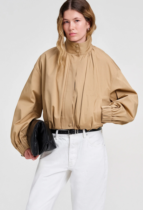 A model wearing a beige high-neck, voluminous long-sleeve jacket with gathered details, paired with white trousers and holding a black clutch bag, against a light gray background.
