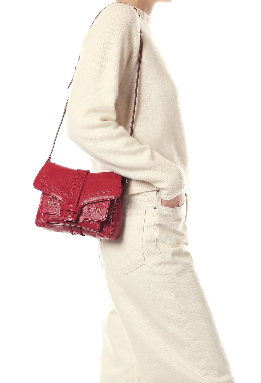 A person wearing a cream ribbed sweater and cream pants carries a small red leather shoulder bag with a flap closure and buckle detail against a plain white background.