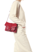 A person wearing a cream ribbed sweater and cream pants carries a small red leather shoulder bag with a flap closure and buckle detail against a plain white background.