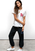 A woman modeling a white graphic t-shirt with red text and black jogger sweatpants with a dark blue graphic, standing against a white wall on a gray concrete floor.