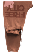 A pair of brown sweatpants featuring a large 'FREE CITY' graphic in dark brown on the upper leg, a pink dove silhouette above it, and a white drawstring at the elastic waistband.