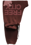 A pair of dark brown sweatpants featuring a large, faded pinkish-brown graphic on the upper leg that reads 'FREE CITY' alongside a silhouette of a dove. The pants have an elasticized ankle cuff with a white drawstring.