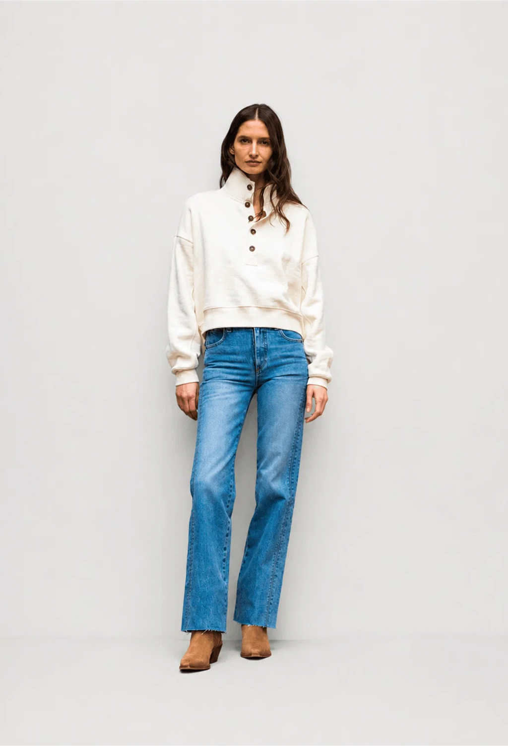 A model stands against a light gray studio background wearing a cream-colored cropped henley sweatshirt with brown buttons and blue straight-leg jeans.