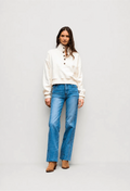 A model stands against a light gray studio background wearing a cream-colored cropped henley sweatshirt with brown buttons and blue straight-leg jeans.