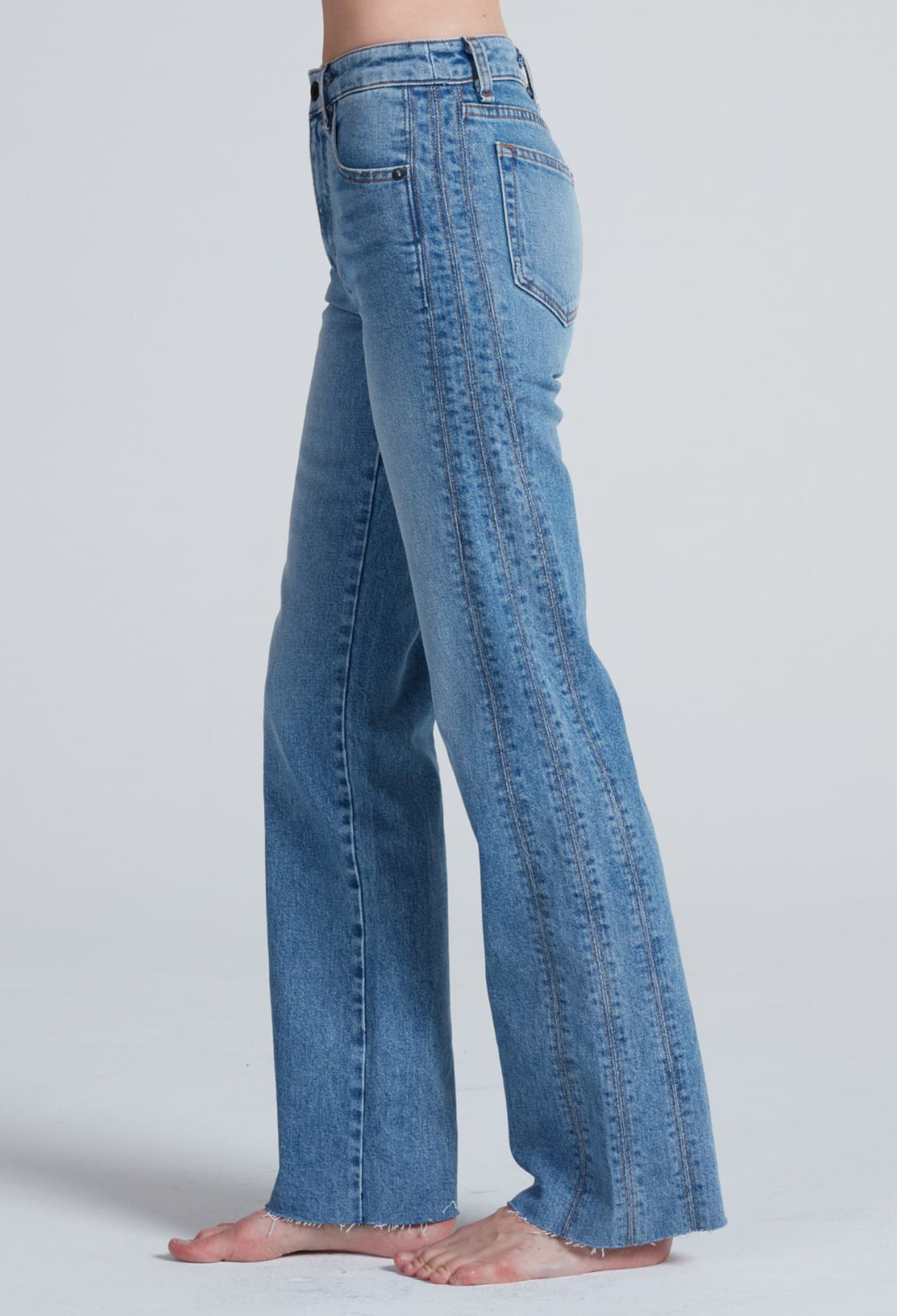 Side view of a person wearing high-waisted, wide-leg blue denim jeans, shown barefoot against a light gray studio background.