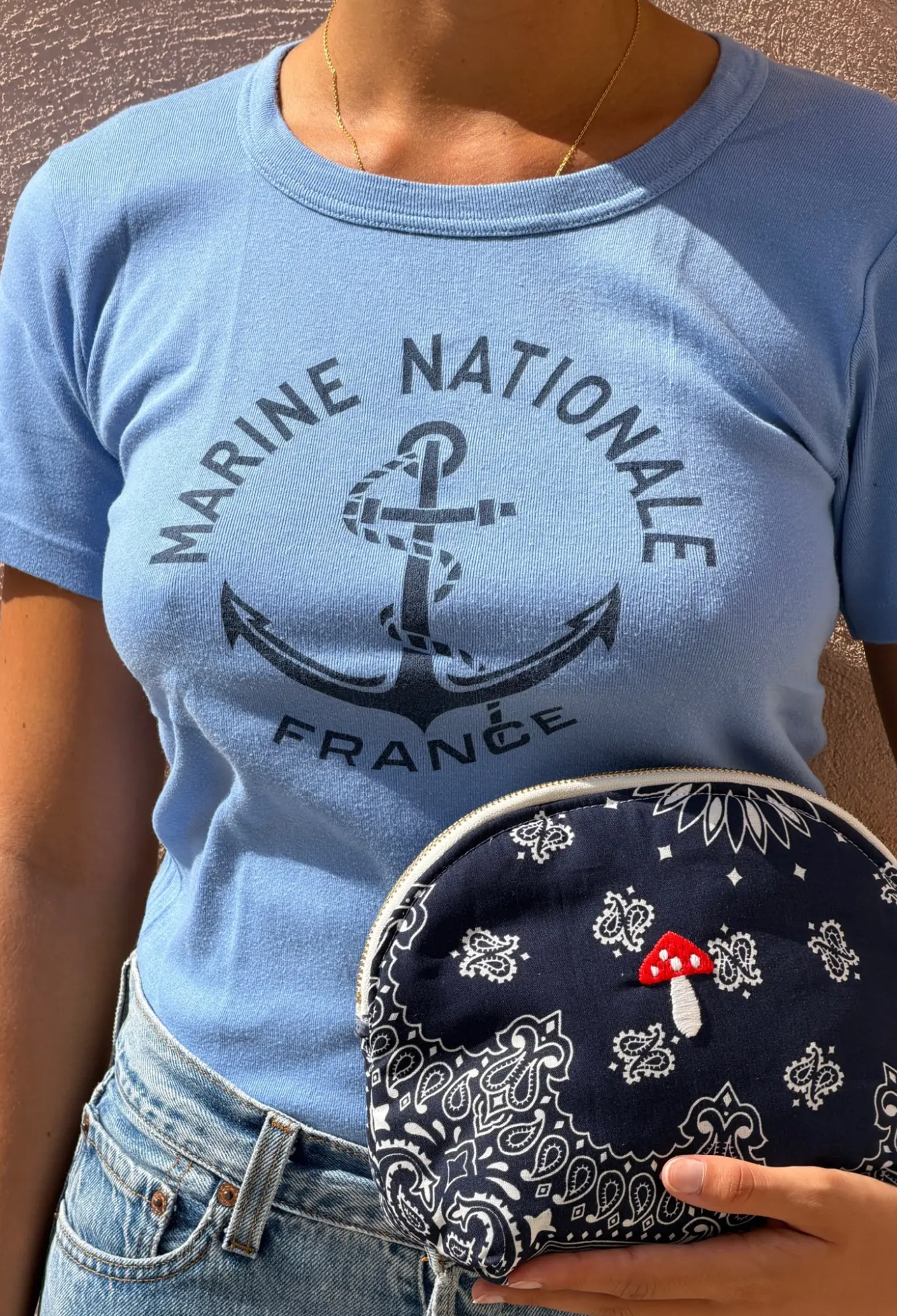 A person wearing a light blue graphic T-shirt holding a navy bandana-patterned pouch with a small red mushroom detail.
