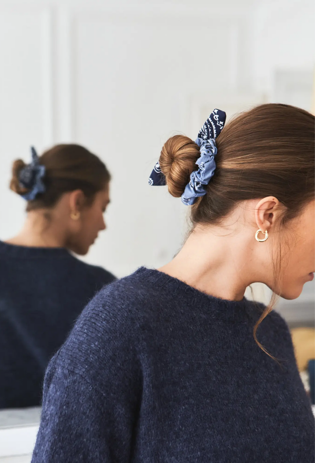 A woman wearing a blue patterned fabric scrunchie in her hair, shown from the side with her reflection visible in a mirror behind her. She is wearing a dark navy knit sweater and a small gold hoop earring.