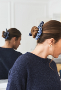 A woman wearing a blue patterned fabric scrunchie in her hair, shown from the side with her reflection visible in a mirror behind her. She is wearing a dark navy knit sweater and a small gold hoop earring.
