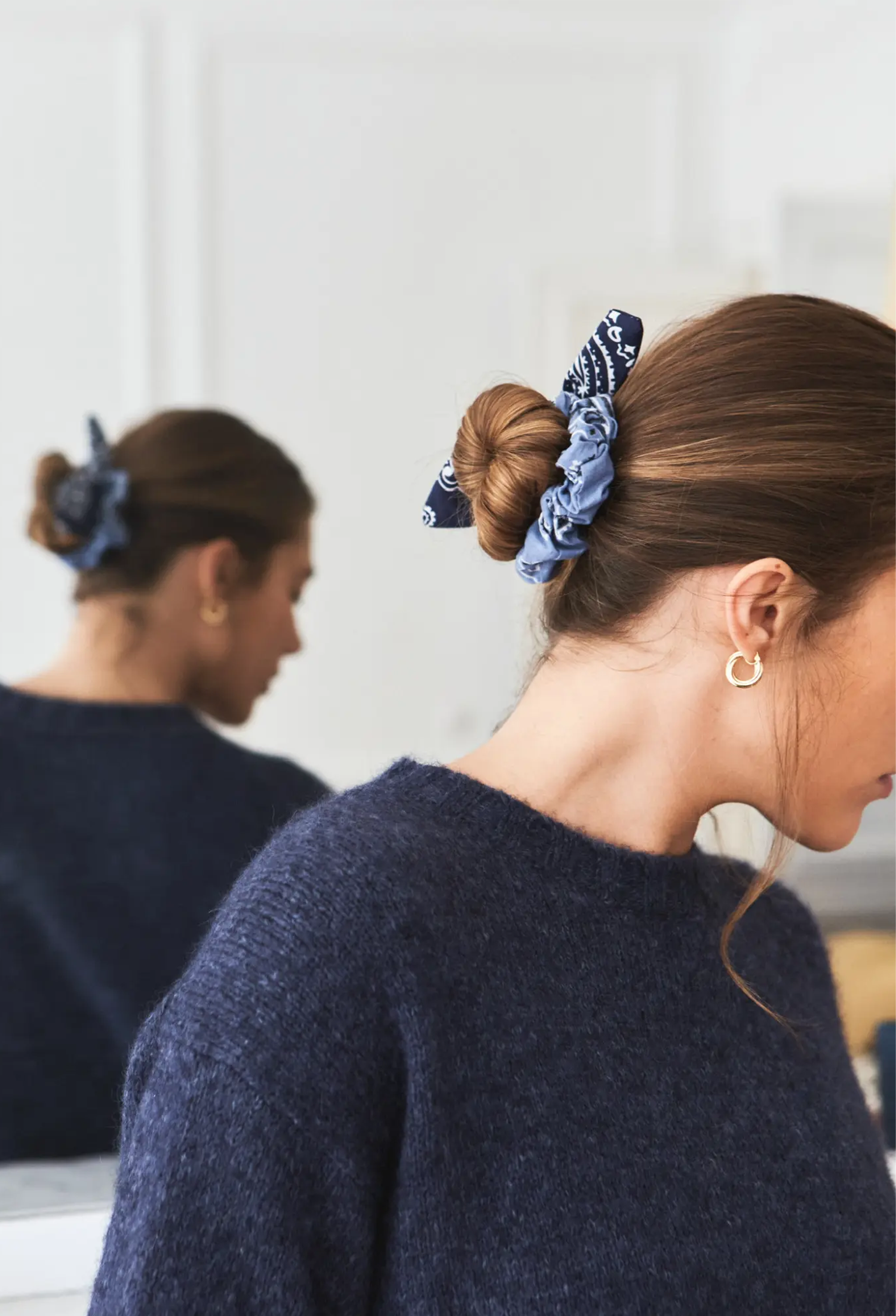 A woman wearing a blue patterned fabric scrunchie in her hair, shown from the side with her reflection visible in a mirror behind her. She is wearing a dark navy knit sweater and a small gold hoop earring.