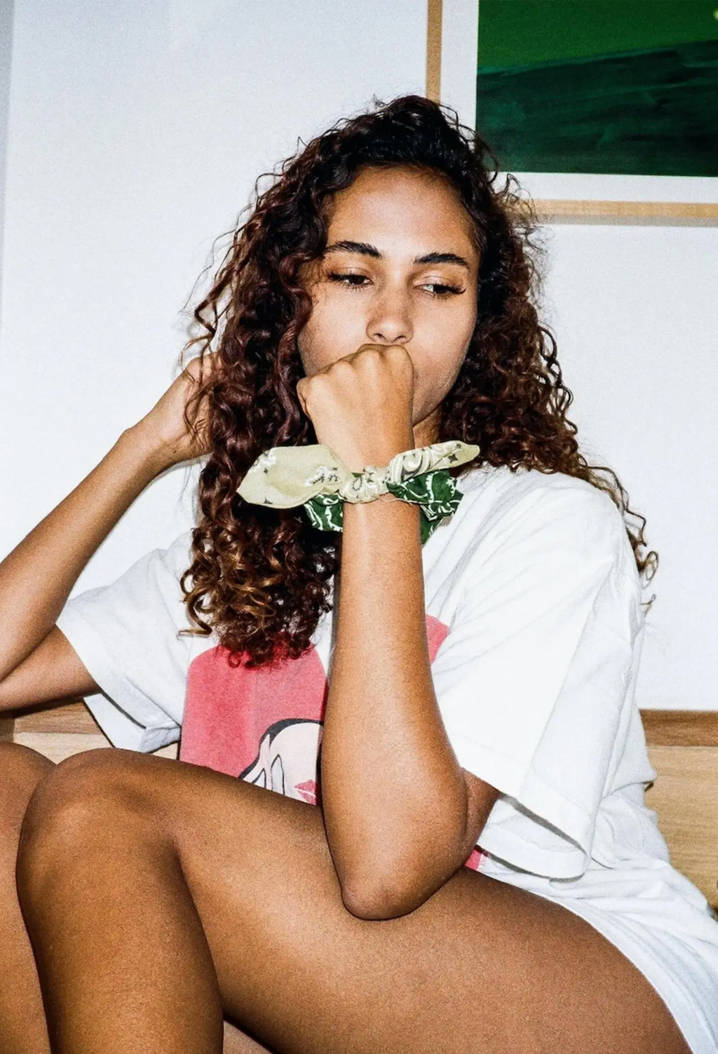 A woman with curly brown hair sits indoors wearing a white graphic T-shirt and a light green and white patterned fabric scrunchie on her wrist.