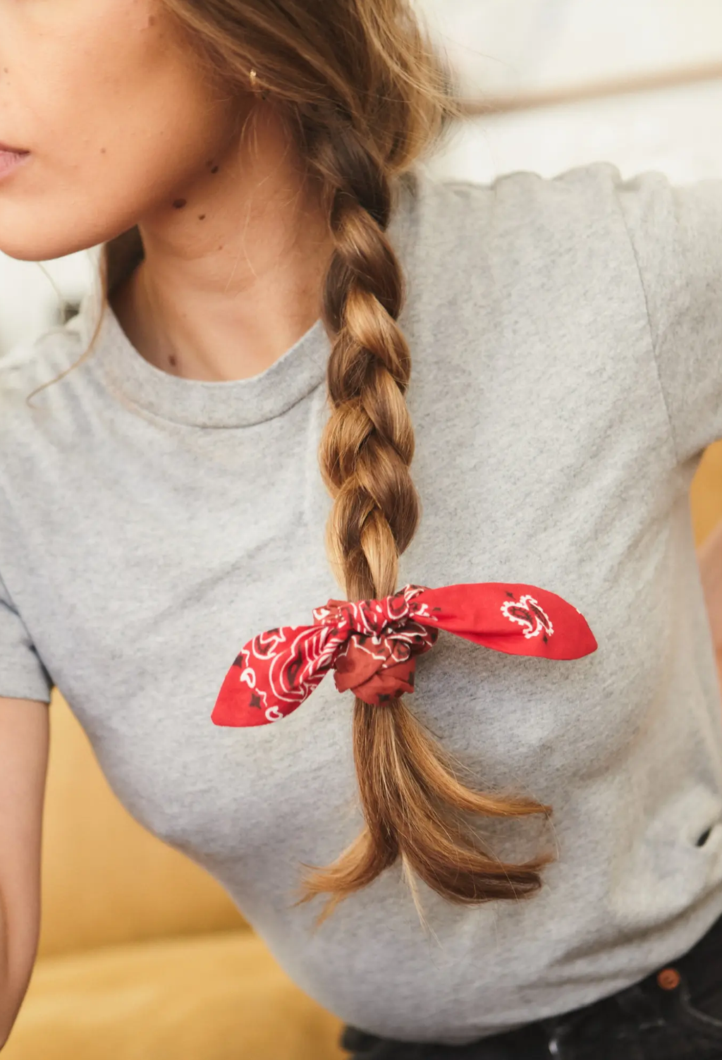 A close-up lifestyle shot of a woman with a long brown braid secured with a red bandana scrunchie, wearing a gray t-shirt.