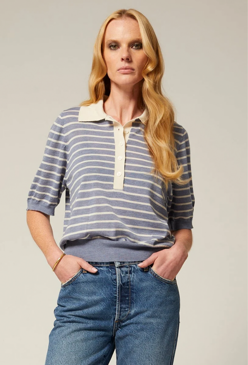 A woman modeling a blue and white striped knit polo shirt with a cream collar and placket, paired with blue jeans, against a solid light gray background.