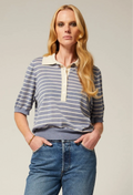 A woman modeling a blue and white striped knit polo shirt with a cream collar and placket, paired with blue jeans, against a solid light gray background.