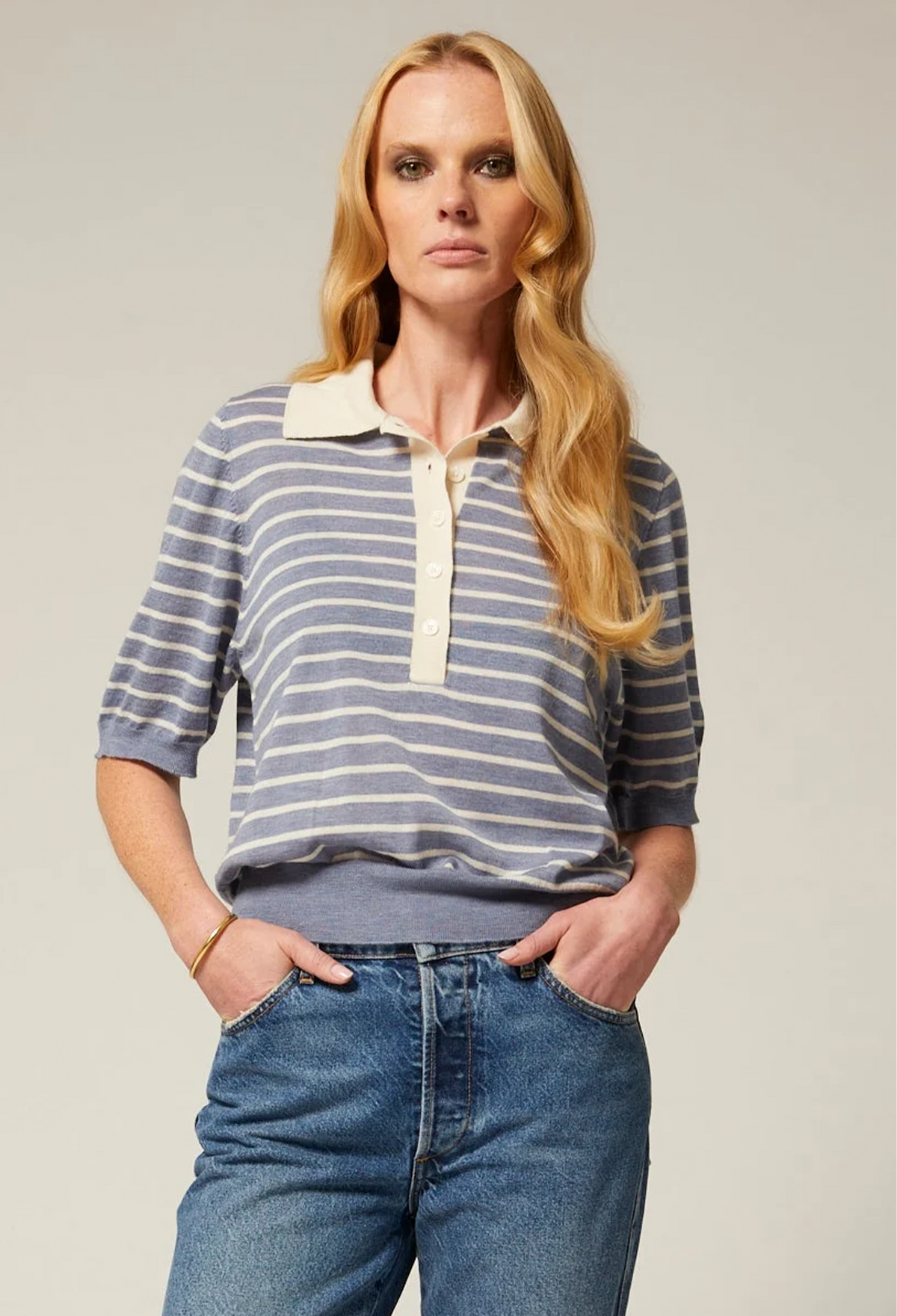 A woman modeling a blue and white striped knit polo shirt with a cream collar and placket, paired with blue jeans, against a solid light gray background.