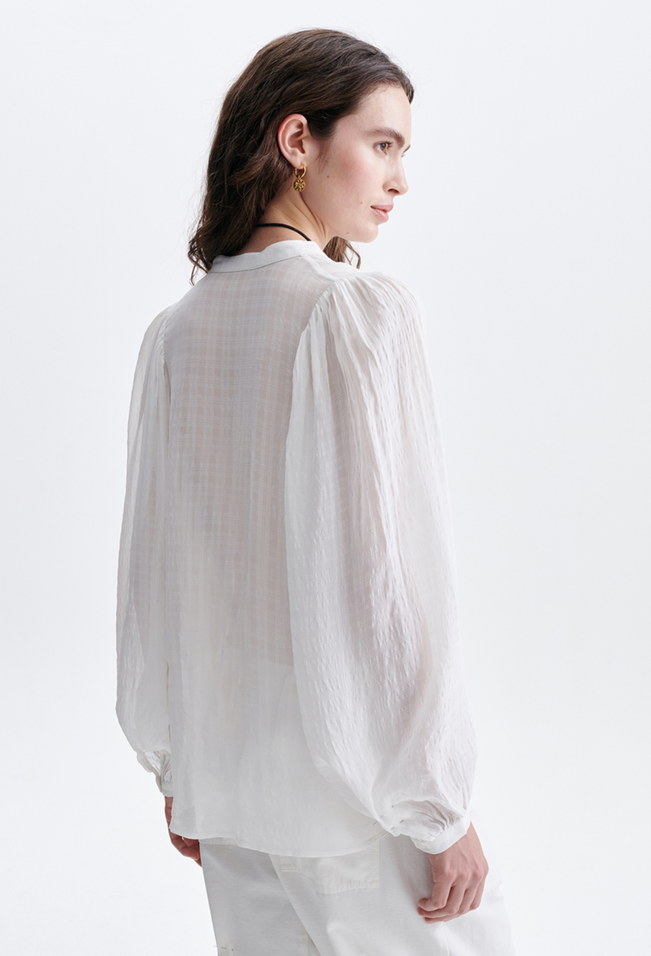 A woman shown from the back wearing a loose white long-sleeve blouse with a sheer, crinkled texture and gathered cuffs, paired with white trousers against a plain white studio background.