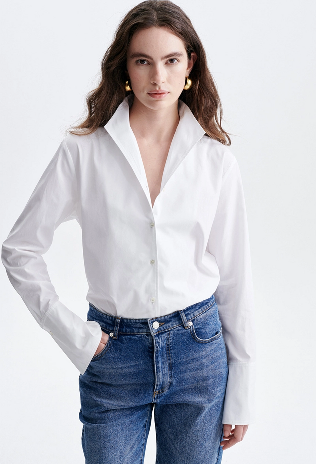 A woman modeling a white long-sleeve button-up shirt tucked into blue jeans, photographed against a clean light gray studio background.