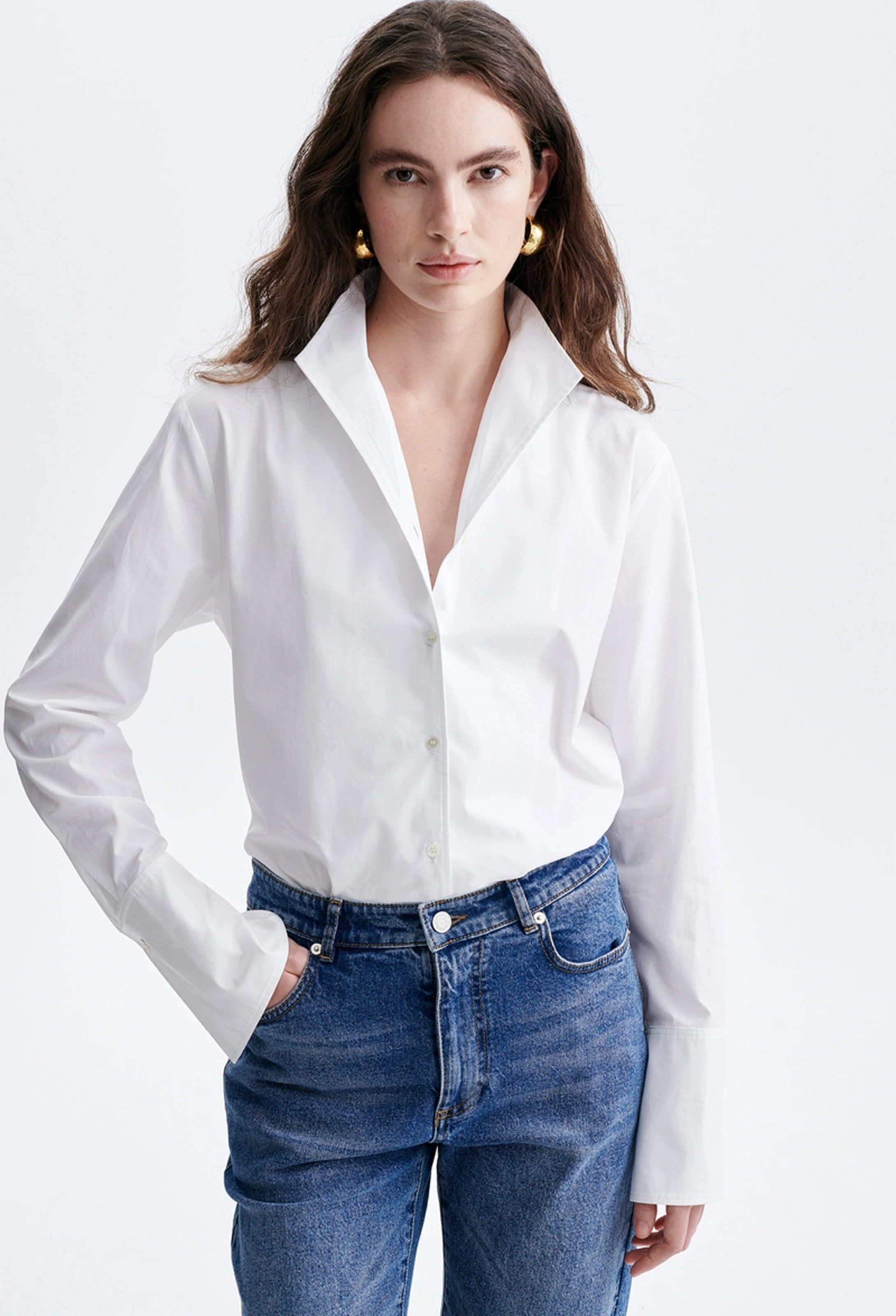A woman modeling a white long-sleeve button-up shirt tucked into blue jeans, photographed against a clean light gray studio background.