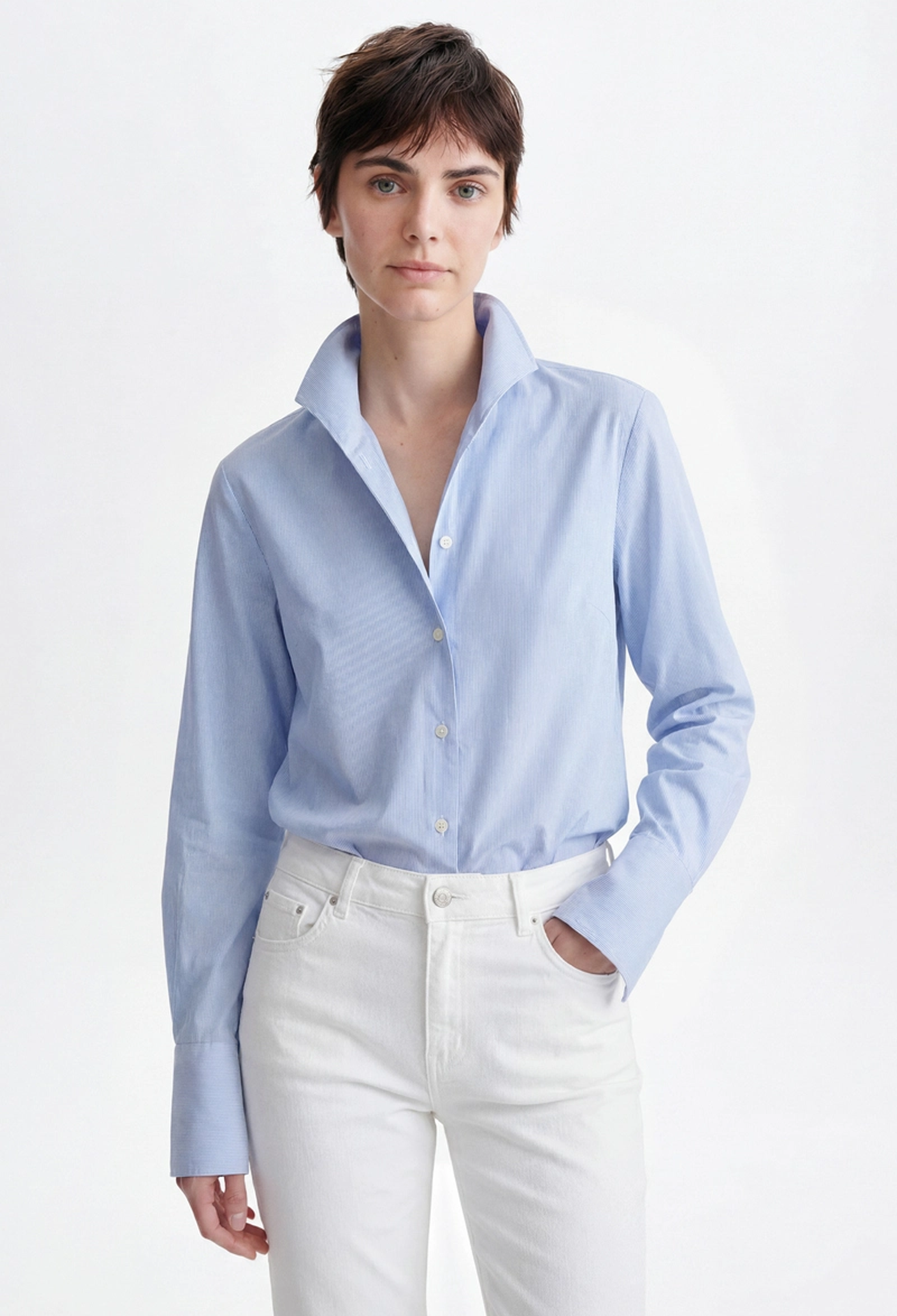 A woman modeling a light blue long-sleeve button-up shirt with white pants, photographed against a clean white studio background.
