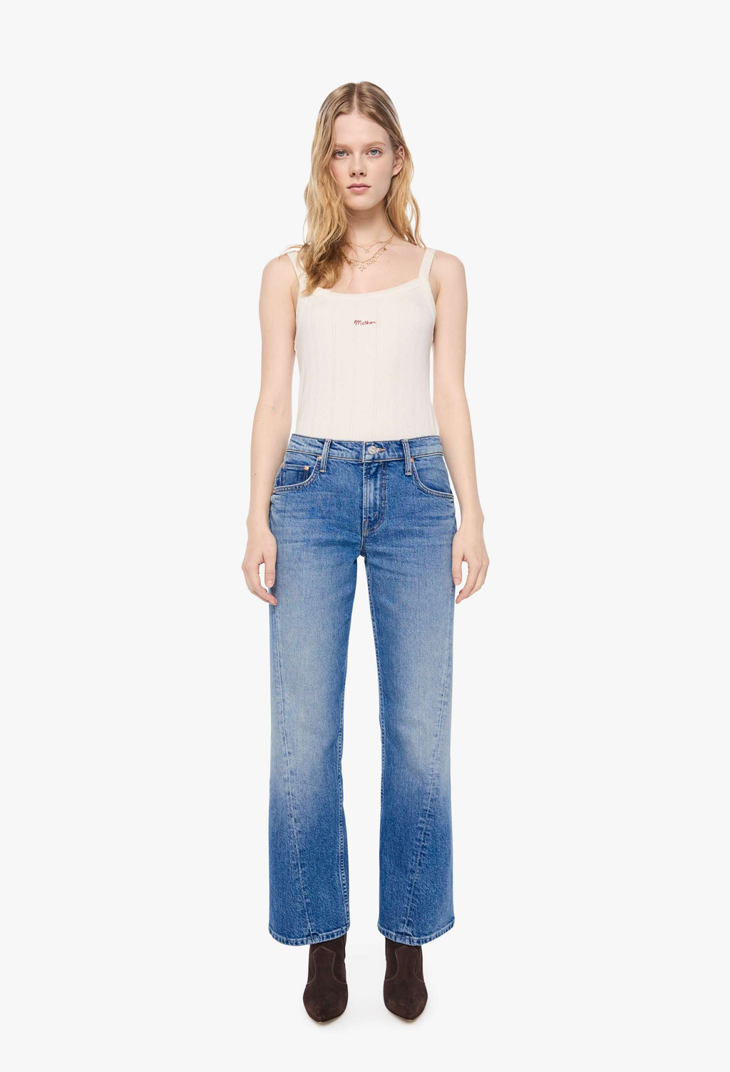 A model stands against a plain white studio background wearing a cream-colored ribbed tank top, blue wide-leg jeans with a faded wash, and dark brown ankle boots.