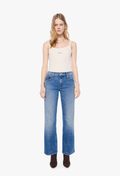 A model stands against a plain white studio background wearing a cream-colored ribbed tank top, blue wide-leg jeans with a faded wash, and dark brown ankle boots.