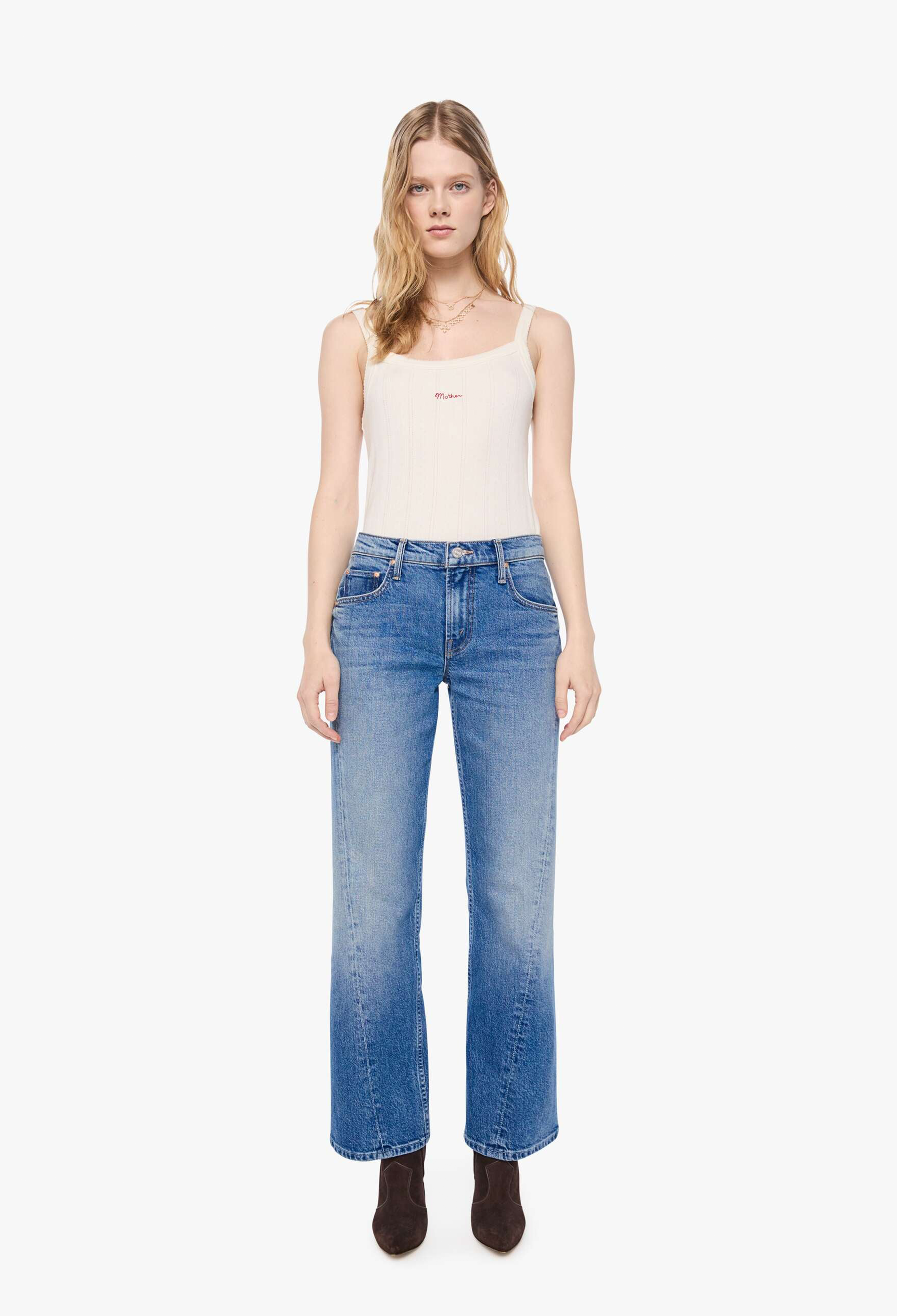 A model stands against a plain white studio background wearing a cream-colored ribbed tank top, blue wide-leg jeans with a faded wash, and dark brown ankle boots.