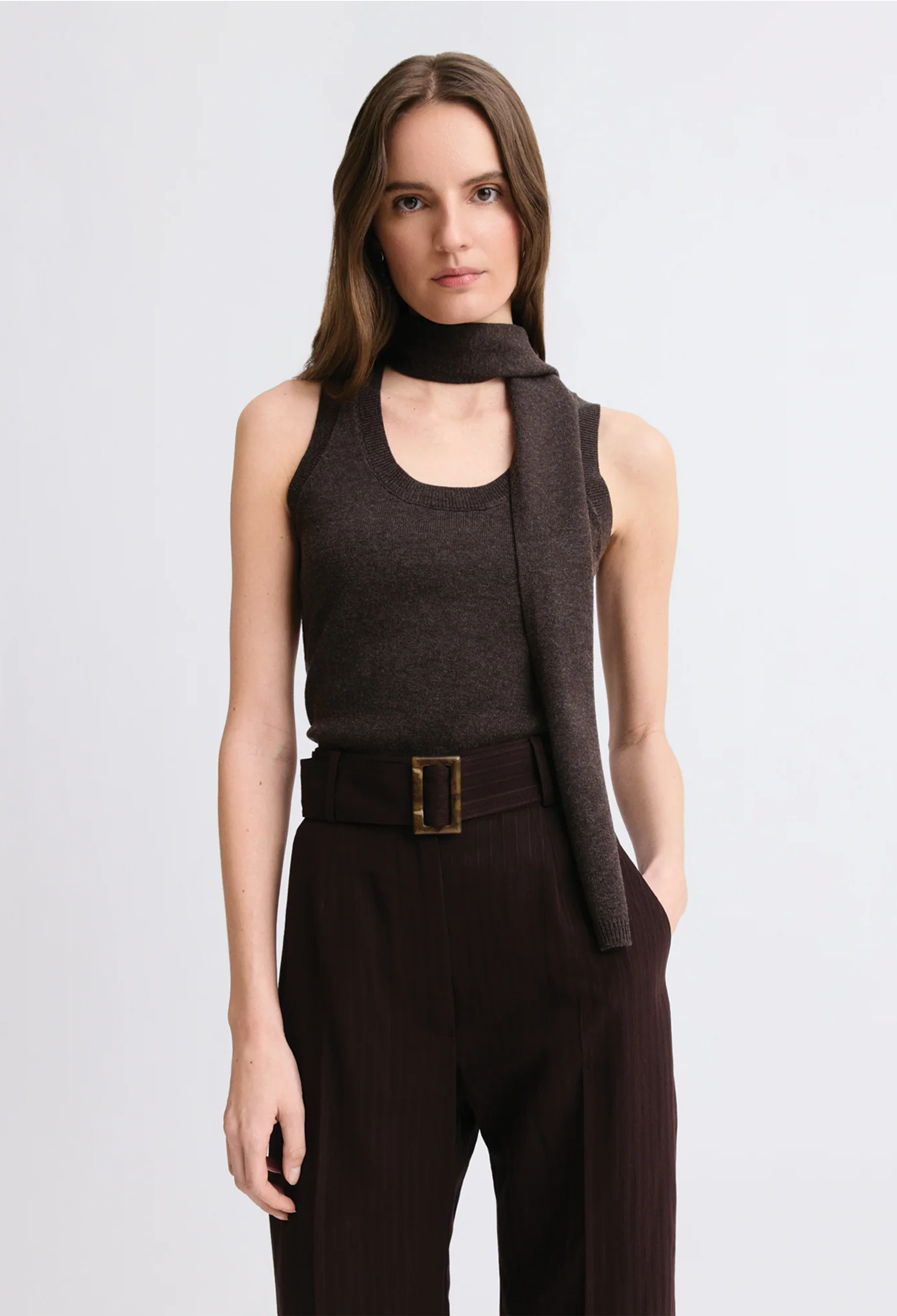 A model wearing a dark brown sleeveless knit top with a draped scarf detail, paired with dark brown trousers and a matching belt with a gold buckle, against a light gray studio background.