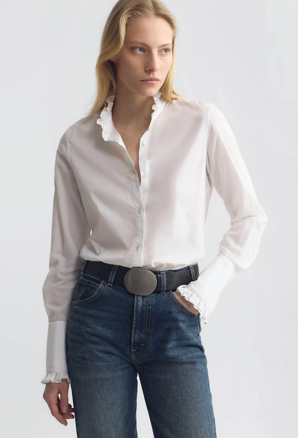 A woman wearing a white long-sleeve button-up blouse with ruffled trim at the collar and cuffs, tucked into dark blue jeans with a black belt, against a light gray studio background.