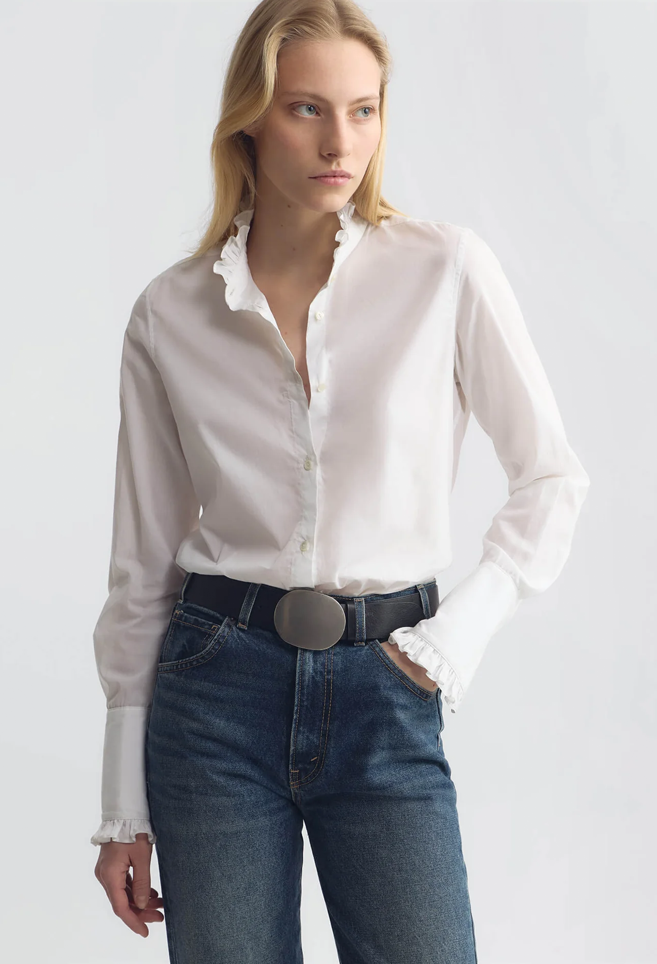 A woman wearing a white long-sleeve button-up blouse with ruffled trim at the collar and cuffs, tucked into dark blue jeans with a black belt, against a light gray studio background.