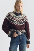 A woman wearing a fuzzy knit Fair Isle sweater with a dark brown base, cream and gray geometric patterns, paired with blue jeans and a black belt, against a light gray studio background.