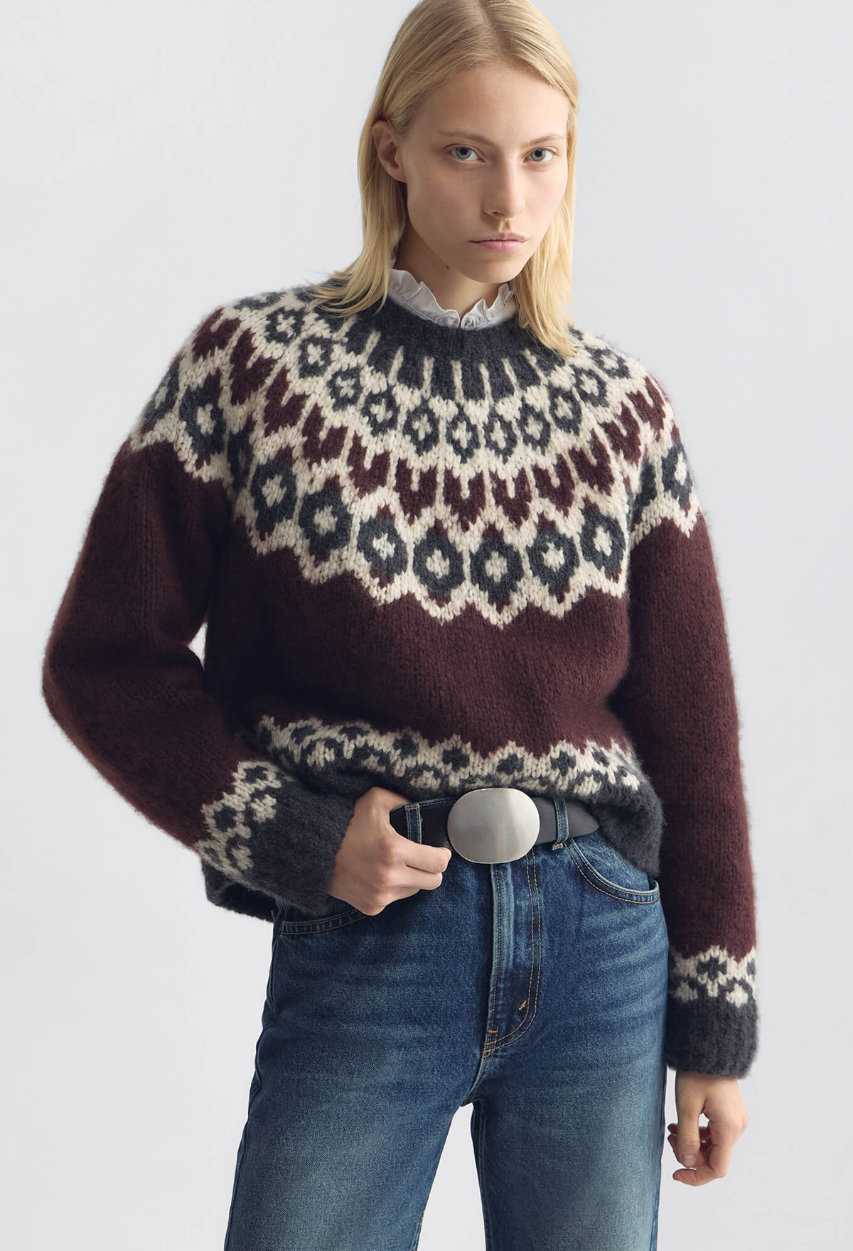 A woman wearing a fuzzy knit Fair Isle sweater with a dark brown base, cream and gray geometric patterns, paired with blue jeans and a black belt, against a light gray studio background.