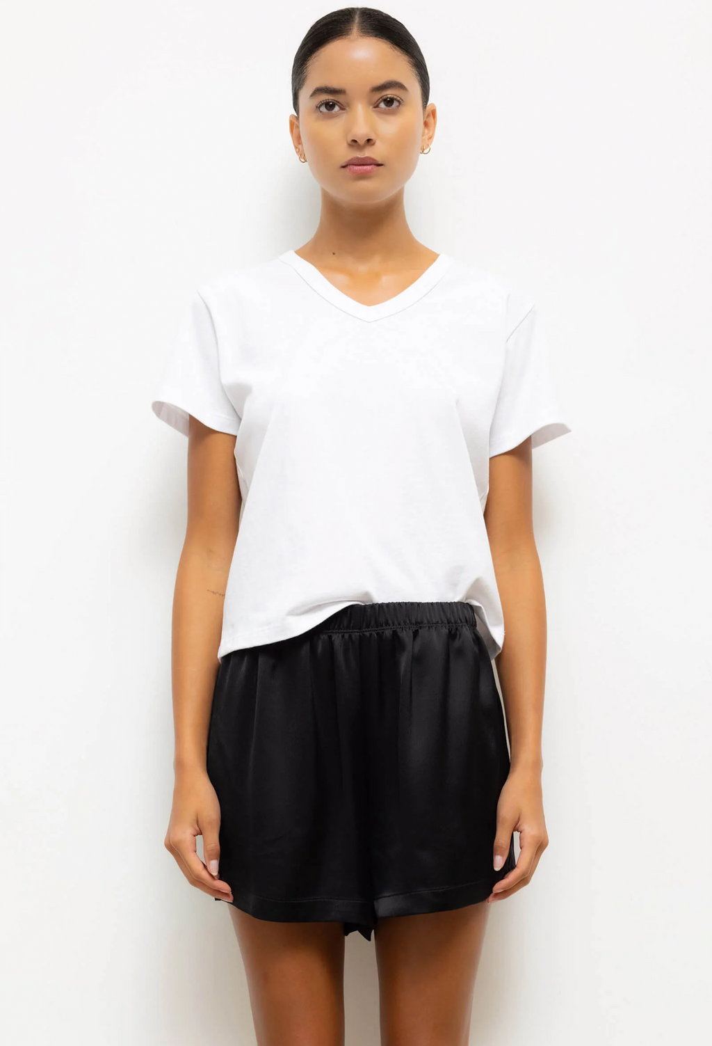 A woman modeling a white short-sleeve V-neck t-shirt and black satin shorts against a plain white background.