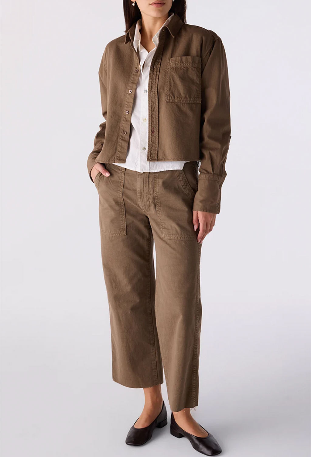 A model wearing a brown cropped button-up shirt jacket, matching brown cropped pants, a white collared shirt, and dark brown shoes, photographed against a light gray studio background.