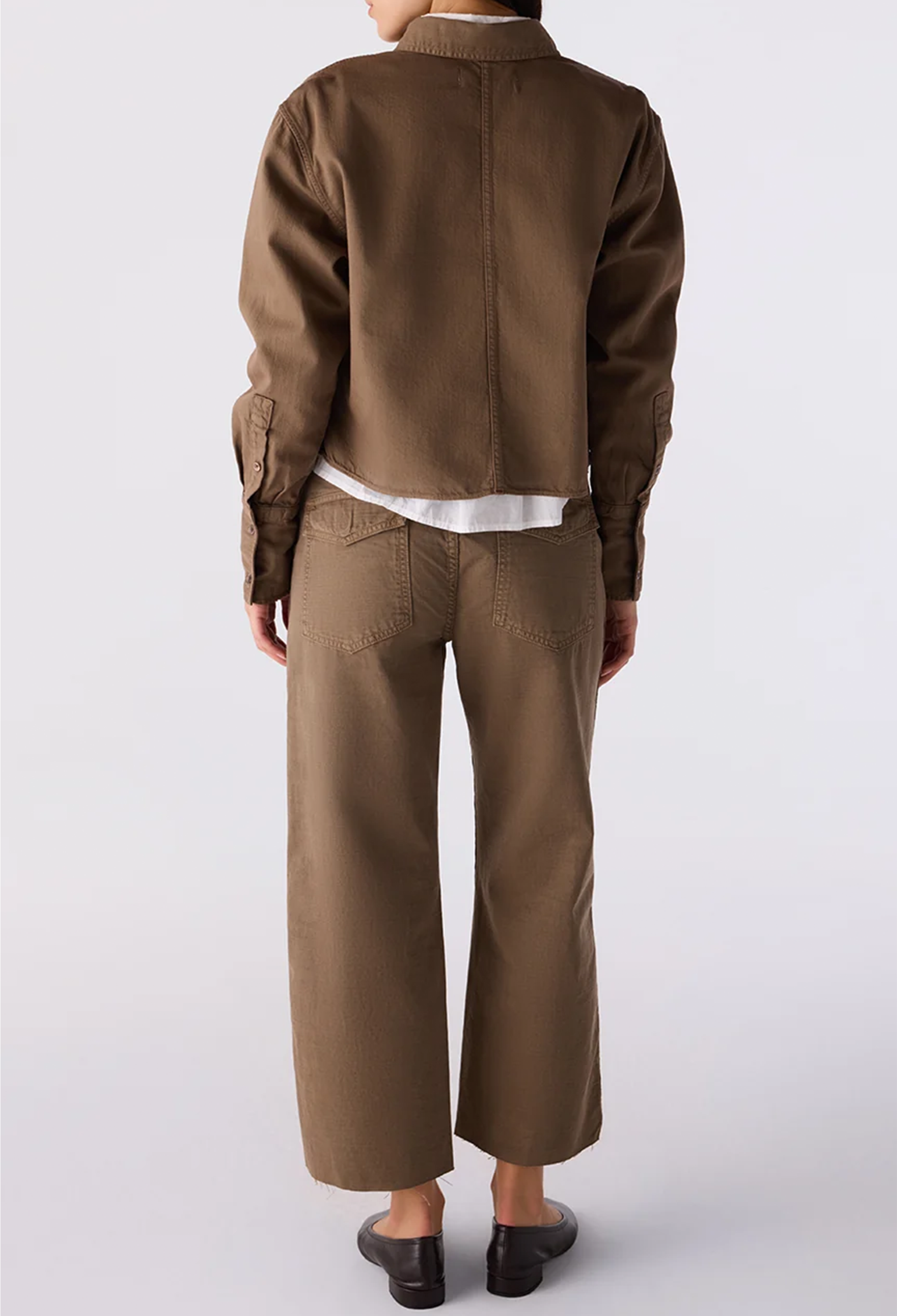 A model shown from the back wearing a brown cropped utility jacket, matching brown wide-leg cropped pants, a white undershirt, and black shoes against a light gray studio background.