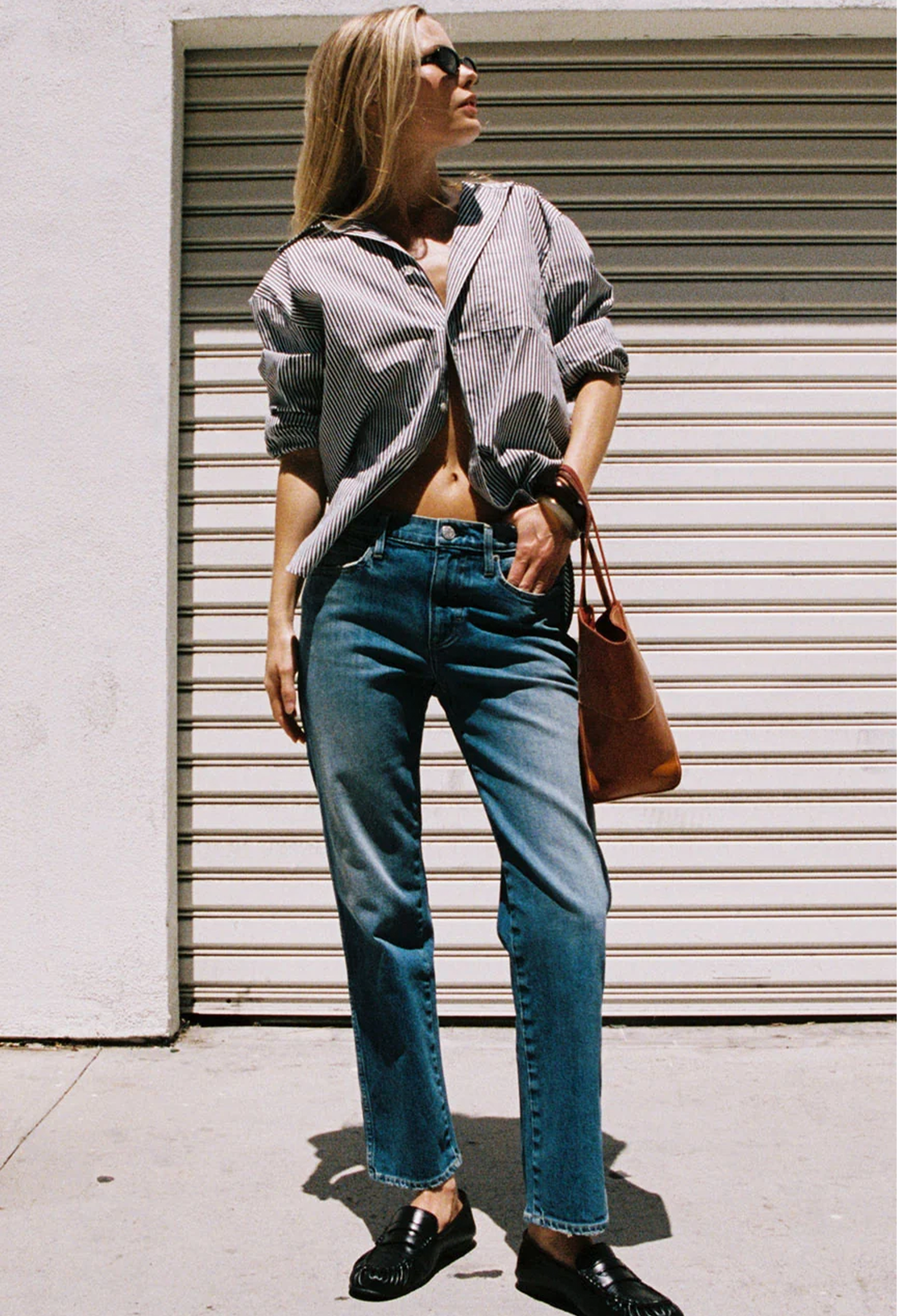 A woman stands outdoors in front of a closed metal roll-up door, wearing an oversized gray-and-white striped button-up shirt tied at the waist, blue jeans, black loafers, and carrying a brown leather tote bag.