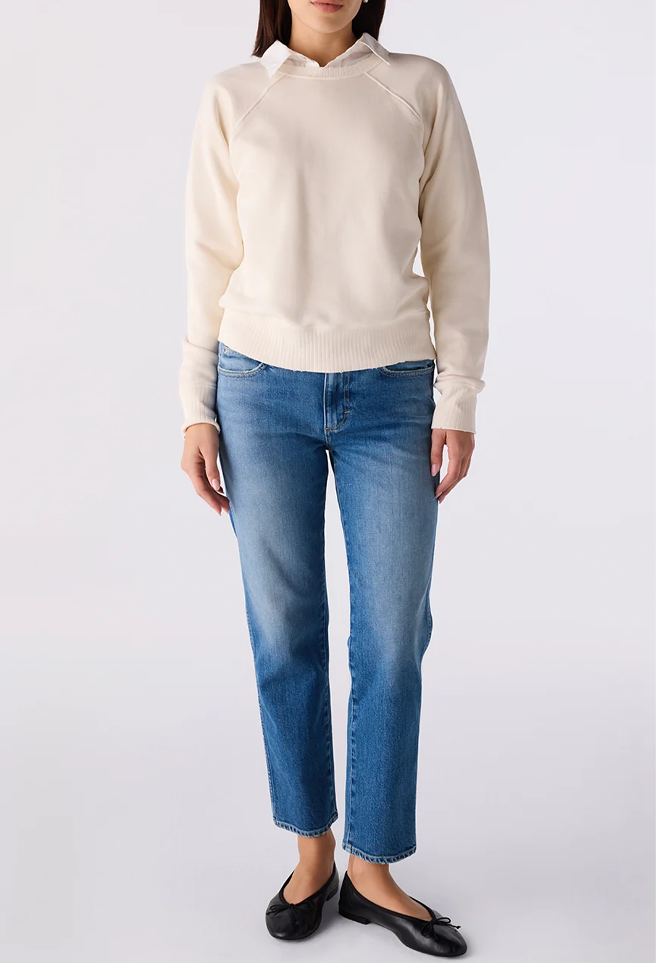 A woman modeling a cream-colored knit sweater with raglan sleeves and a mock neck, paired with blue cropped jeans and black flat shoes, against a light gray studio background.