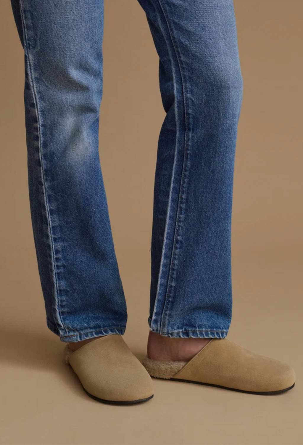 Close-up of a person's legs wearing blue denim jeans and beige suede mule slip-on shoes against a solid tan background.