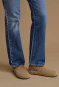 Close-up of a person's legs wearing blue denim jeans and beige suede mule slip-on shoes against a solid tan background.